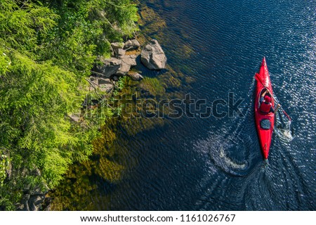 River Kayaker Aerial View. Caucasian Sportsman in the Red Kayak Paddling on the Scenic River Along the Shore.