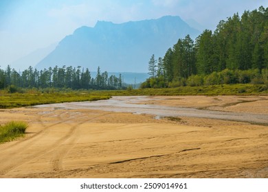 A river meanders through a sandy valley, surrounded by green trees and a towering mountain range in the background under a clear sky.  - Powered by Shutterstock