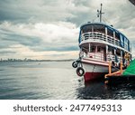 A river navigation boat seen from its bow anchored at a pier in a small port with the city of Manaus in the background on a cloudy day.