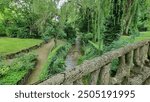 A river view from an old stone bridge, showing the flowing water beneath. The scene includes lush greenery along the riverbanks and the stone railings of the bridge in the foreground.