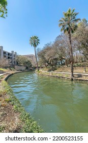 The River Walk At San Antonio Texas With Winding Canal Flanked By Trails. Scenic Nature Views And Buildings Can Also Be Seen Against Blue Sky On This Sunny Day.