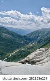 A river winds through a forested alpine valley below the Rhone Glacier, with the switchback road of the Furka Pass visible carving through the green mountainside in Switzerland.