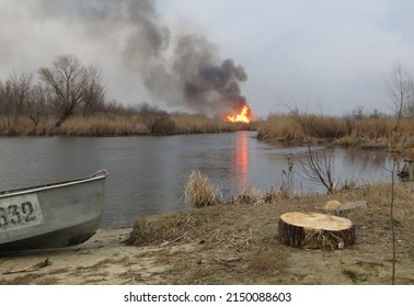 Riverside with boat and stump against the background of burning forest and smoke and reeds and river under overcast sky 