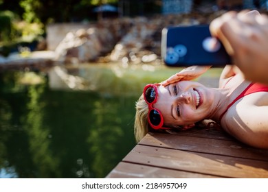 Rlaxed woman relaxing on the pier, lying and taking selfie near the natural lake in summer