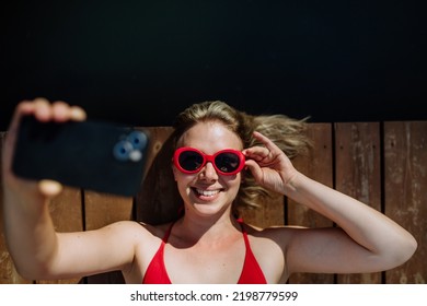 Rlaxed woman taking selfie when lying on a pier by natural lake in summer