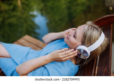 Rlaxed woman wearing headphones listening to music lying on a pier by natureal lake in summer