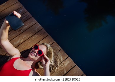Rlaxed woman wearing headphones listening to music and taking selfie when lying on a pier by natural lake in summer