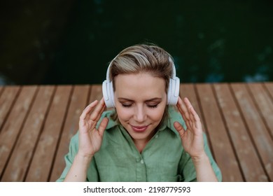 Rlaxed woman wearing headphones listening to music sitting on a pier by natureal lake in summer