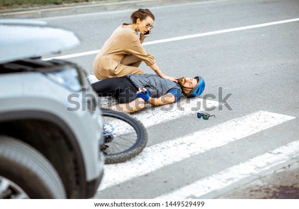 Road accident with injured cyclist lying on the\
pedestrian crossing near the broken bicycle, worried woman driver\
calling and checking men\'s\
pulse