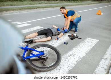 Road accident with injured cyclist on the pedestrian crossing with passerby pedestrian providing first aid fixing men's head
