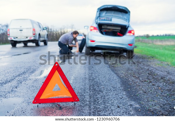 road accident sign\
on the background man replaces flat tyre on road. Car tire leak\
because of nail pounding. 