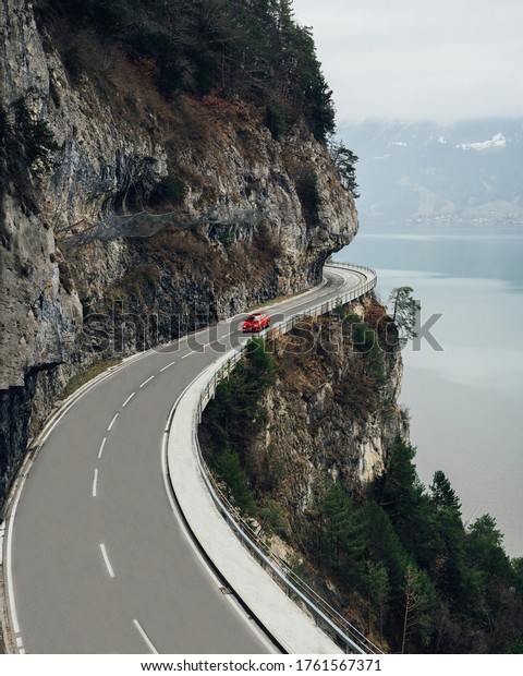Road with beautiful views of the lake and mountains\
of Switzerland. A red car is driving on a winding road near\
Interlaken in the Swiss\
Alps.