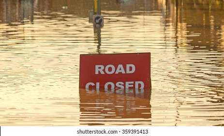 A 'Road Closed' Sign Partially Covered In Flood Water Lit By The Evening Sun