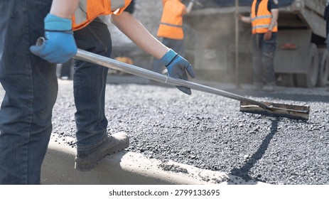 Road construction worker leveling fresh hot asphalt next to a concrete curb with a team in the background