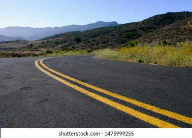 A road curves off into the hills below a blue sky.  The hills are Mediterranean climate.  The road has a double yellow line.