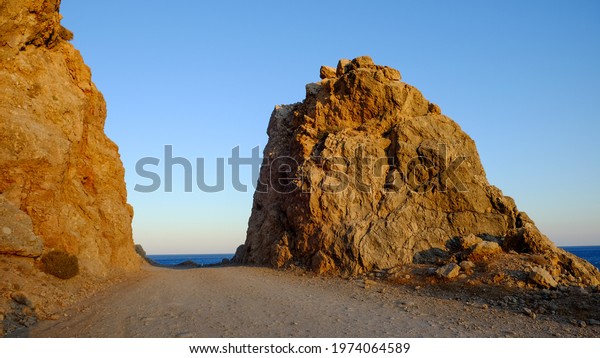 The road cut in the rock deviding it in two\
cliffs lit by the sun, sunset sky on the background metting the sea\
on the horizon line, Crete,\
Greece