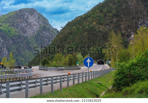 Road fork on\
a highway before entering a tunnel in the mountains in northern\
Italy, road sign in green grass on the side of the road, cloudy\
sky, natural Europe travel\
background