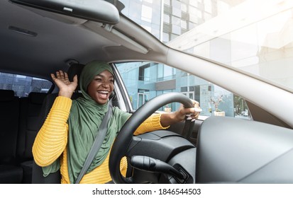 Road Fun. Joyful African Muslim Woman In Hijab Dancing In Car, Singing While Driving Her New Vehicle, Cheerful Black Islamic Lady In Headscarf Enjoying Road Drive In City, Low Angle View