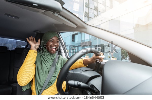 Road Fun. Joyful African Muslim Woman In Hijab\
Dancing In Car, Singing While Driving Her New Vehicle, Cheerful\
Black Islamic Lady In Headscarf Enjoying Road Drive In City, Low\
Angle View
