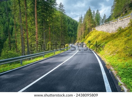 Road in green forest at sunset in summer. Dolomites, Italy. Beautiful mountain roadway, tress, grass, high rocks, blue sky with clouds. Landscape with empty highway through the woods in spring. Travel