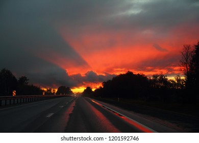Road to hell - evening empty suburban highway after rain on the background of a reflection blazing sunset over the forest - bright orange sun with rays into sky shines through the silhouettes of trees