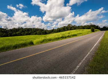 Road Up Hill With Green Grass Field Under White Clouds And Blue Sky.