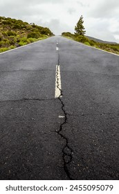 A road with a large crack in it. The road is empty and the sky is cloudy. Scene is somber and desolate