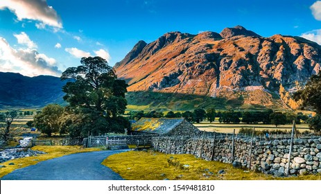 Road leading to picturesque stone cottage house next to a river with Great Langdale mountains and valley in the background at sunset time in the Lake District National Park in North West England, UK.