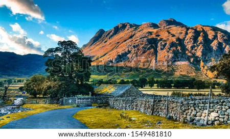 Road leading to picturesque stone cottage house next to a river with Great Langdale mountains and valley in the background at sunset time in the Lake District National Park in North West England, UK.