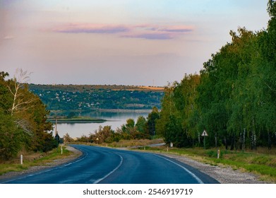 Road Leading to Scenic Lakeside Village at Sunset. A winding road descending towards a peaceful lakeside village with a serene backdrop of trees and pastel skies - Powered by Shutterstock