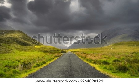 Road leading trough Doolough Valley, between Glenummera and Glencullin mountain ranges illuminated by sunlight with dark dramatic sky, Mayo, Ireland