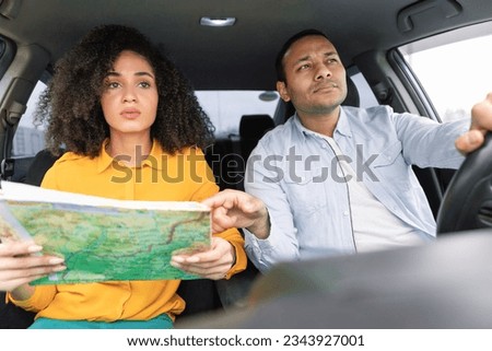 Road Navigation Issue. Perplexed Arab couple riding car holding and pointing at map choosing route and having problems navigating vehicle, sitting in automobile, view from dashboard