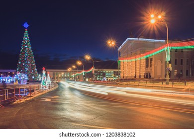 Road at night with the traffic of cars passes by a festive New Year tree and a drama theater