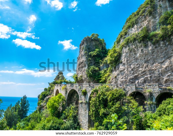 The road over the rocky cliff, right on the\
Mediterranean sea, with luxuriant vegetation all around. - Amalfi\
Coastline road, Naples,\
Italy.