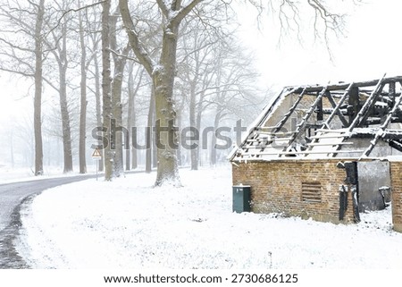 Road passing by burned down barn covered in snow after a blizzard snowstorm with the black wood and construction carcas contrasting against the white hazy winter landscape behind