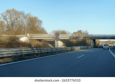 A road runs parallel to an overpass marking a speed limit of 100 km h. The sun shines brightly, illuminating the trees lining the roadside on a crisp morning.