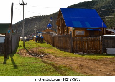 Road In Russian Village With Church And Houses With Blue Roof With Fence In The Evening At Sunset Light. Tractor, Pillars, Green Grass And Mountains Behind