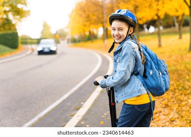 Road to school. Happy kid riding scooter on autumn city street. Child girl with protective helmet and blue backpack on safety bikeway. Back to school concept. Ecological transportation and lifestyle - Powered by Shutterstock