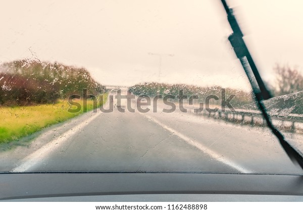 road seen\
through a wet windscreen whilst driving in the rain. One wiper can\
be seen moving across the\
windshield