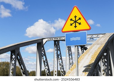 Road sign before a steel bridge, warning of river crossing and potential ice formation. Hazardous bridge passage during winter conditions. Snowflake icon on traffic sign in yellow triangle red border. - Powered by Shutterstock