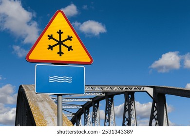 Road sign before a steel bridge, warning of river crossing and potential ice formation. Hazardous bridge passage during winter conditions. Snowflake icon on traffic sign in yellow triangle red border. - Powered by Shutterstock