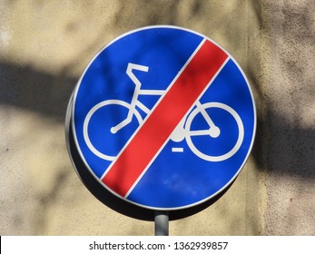 Road Sign “End Of Dedicated Cycle Path”: Round Blue Sign With White Bicycle Symbol And Red Line Strike Through Against A Background Of Blurred Wall In Early Spring, Some Shadows