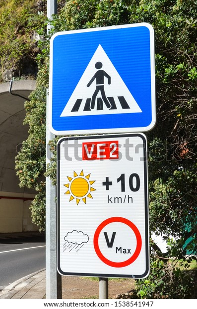 Road signs in Portuguese island Madeira. Blue\
zebra crossing sign. White speed limit sign. The speed restriction\
is dependent on weather, in case of clear weather the limit is plus\
10 kmh than stated.