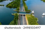 A road stretches across a canal, creating a unique bridge over water. Lush green foliage borders the waterway and a small sailboat drifts peacefully nearby. Veluwemeer Aquaduct in the Netherlands