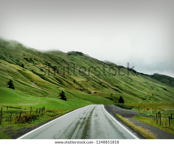 Road\
through the beautiful green scenery in Europe with foggy background\
and mountains. Roadtrip with car into the\
nature