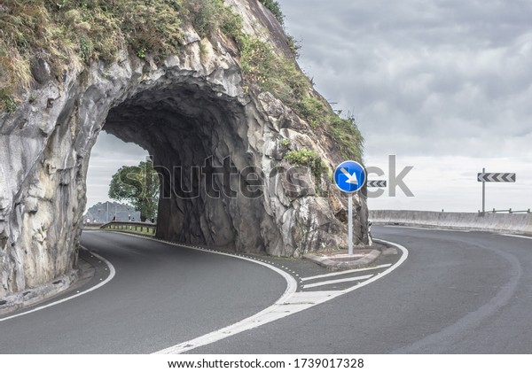 The road through the mountains. Sign of a sharp turn to\
the left on the highway. Road marking. Empty road. Tourism\
industry. Closed borders. Lack of tourists. Stone arch. Road tunnel\
in the rock. 