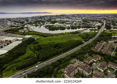 Road in town by a green park and residential area with homes and river. Sunset time. Stunning nature scenery in the background. Galway city, Ireland. Aerial view. City transportation system. - Shutterstock ID 2226124269