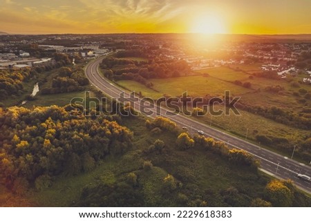 Road in town by a green park and residential area with homes at warm sunset. Sunset time. Stunning nature scenery in the background. Galway city, Ireland. Aerial view. City transportation system.