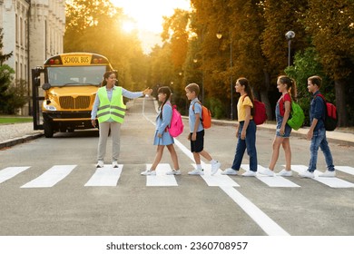 Road Traffic Safety. School Bus Assistant Lady Helping Kids To Cross Road By Crosswalk, African American Attendant Woman In Uniform Guiding Children, Group Of Pupils Walking On Pedestrian Crossing - Powered by Shutterstock