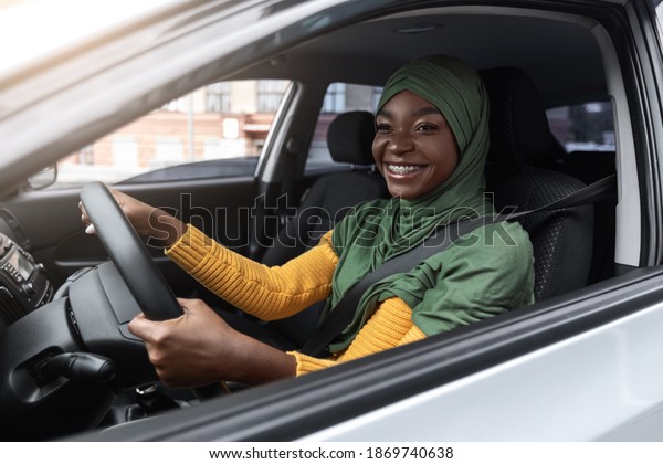 Road Trip. Black Muslim Driver Woman In Hijab\
Enjoying Driving Car In City, Holding Steering Wheel And Smiling\
Through Window, Positive Religious Female With Dental Braces Riding\
Modern Vehicle